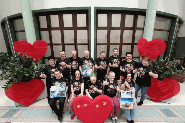 Mieszkańcy w koszulkach WOŚP na holu domu trzymaja obrazy na aukcję.