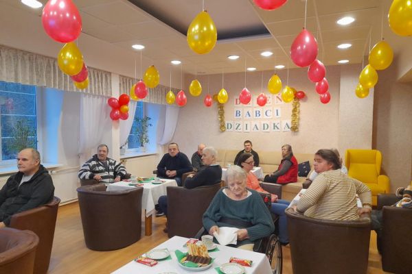 Mieszkańcy przy stolikach z poczęstunkiem i muzyce obchodzą Dzień Babci i Dziadka.