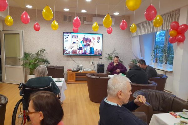 Mieszkańcy przy stolikach z poczęstunkiem i muzyce obchodzą Dzień Babci i Dziadka.