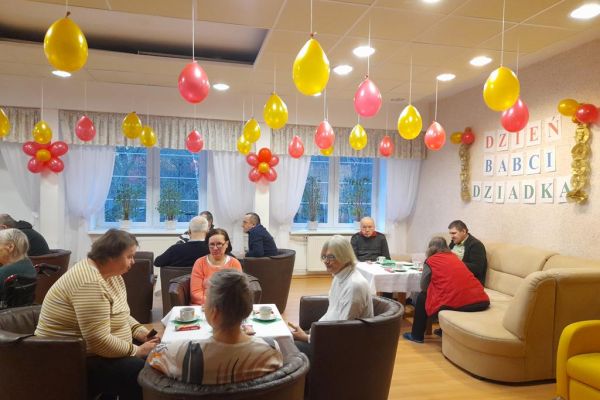 Mieszkańcy przy stolikach z poczęstunkiem i muzyce obchodzą Dzień Babci i Dziadka.