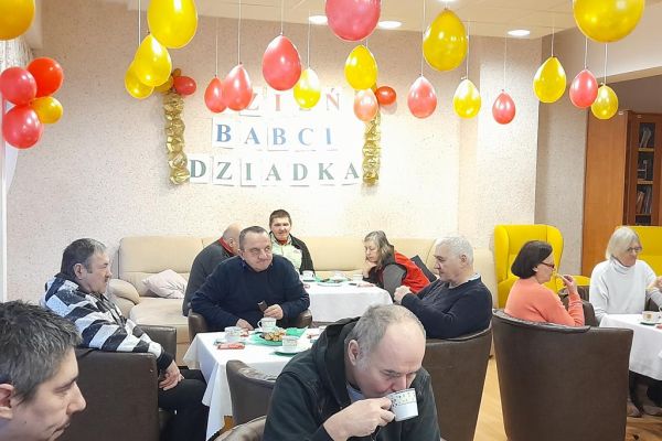Mieszkańcy przy stolikach z poczęstunkiem i muzyce obchodzą Dzień Babci i Dziadka.