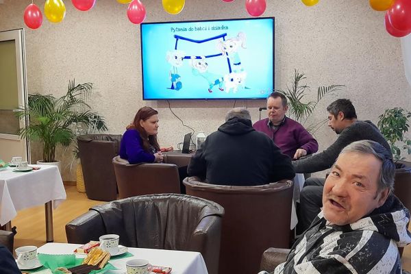 Mieszkańcy przy stolikach z poczęstunkiem i muzyce obchodzą Dzień Babci i Dziadka.