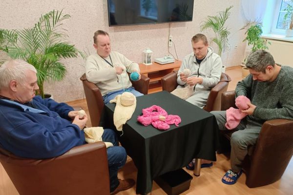 Mieszkańcy uczestniczą w zajęciach z ergoterapii - siedzą przy stolikach, zwijają włóczkę, niektórzy dziergają, zwijają kłębki.