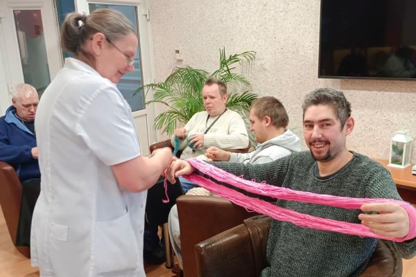 Mieszkańcy uczestniczą w zajęciach z ergoterapii - siedzą przy stolikach, zwijają włóczkę, niektórzy dziergają, zwijają kłębki.