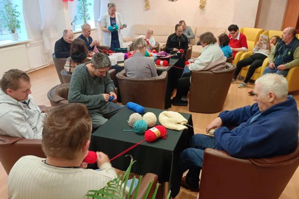 Mieszkańcy uczestniczą w zajęciach z ergoterapii - siedzą przy stolikach, zwijają włóczkę, niektórzy dziergają, zwijają kłębki.