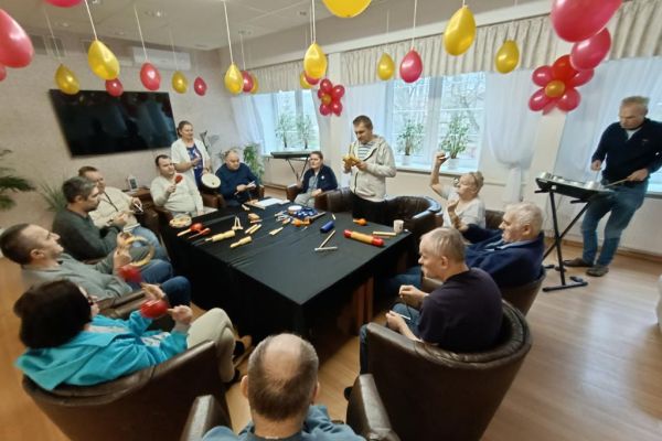 Mieszkańcy ćwiczą i wystukują rytmy na róznych instrumwentach siedząc w fotelikach przy stole wyłożonym instrumentami.