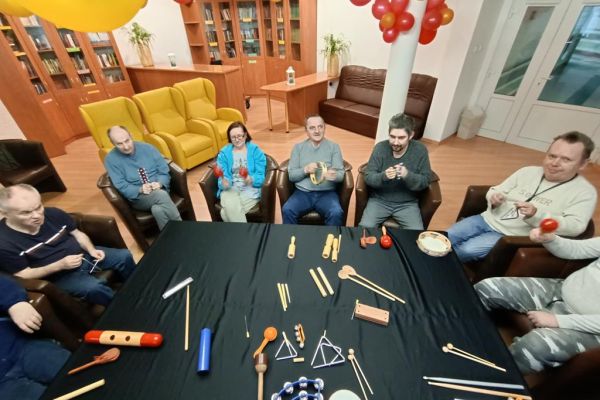 Mieszkańcy ćwiczą i wystukują rytmy na róznych instrumwentach siedząc w fotelikach przy stole wyłożonym instrumentami.