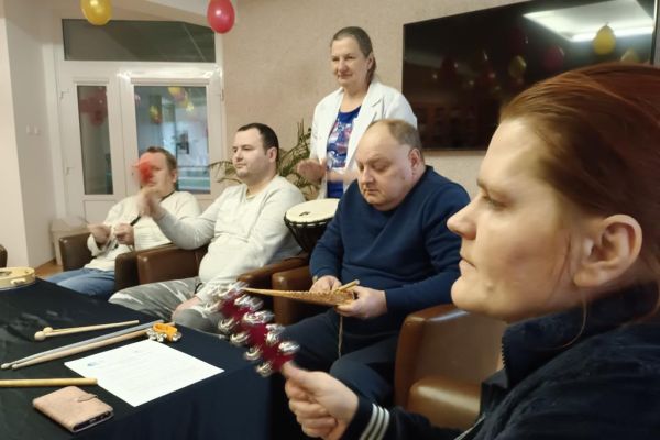 Mieszkańcy ćwiczą i wystukują rytmy na róznych instrumwentach siedząc w fotelikach przy stole wyłożonym instrumentami.