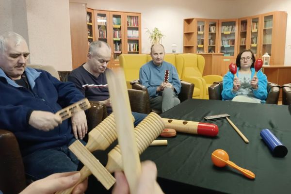 Mieszkańcy ćwiczą i wystukują rytmy na róznych instrumwentach siedząc w fotelikach przy stole wyłożonym instrumentami.