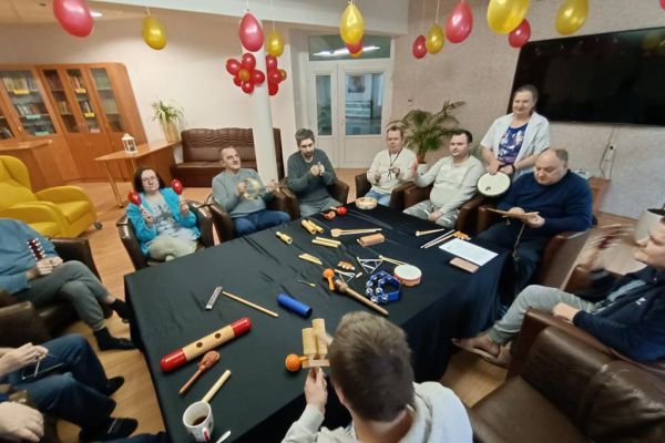Mieszkańcy ćwiczą i wystukują rytmy na róznych instrumwentach siedząc w fotelikach przy stole wyłożonym instrumentami.