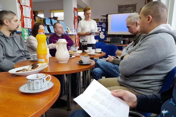 Mieszkańcy siedzą w bibliotece przy stolikach i wysłuchują Pani prowadzącej.
