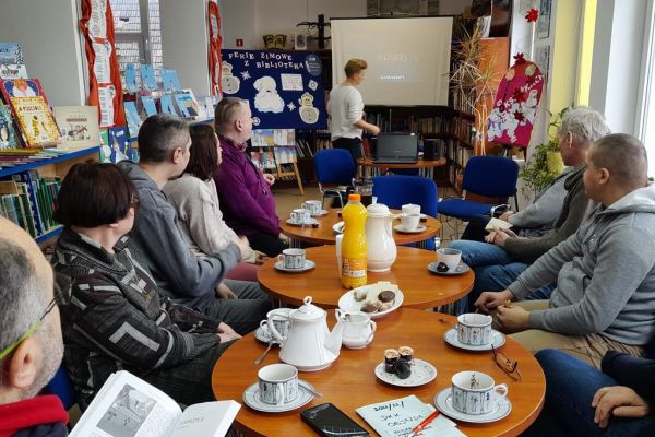 Mieszkańcy siedzą w bibliotece przy stolikach i wysłuchują Pani prowadzącej.