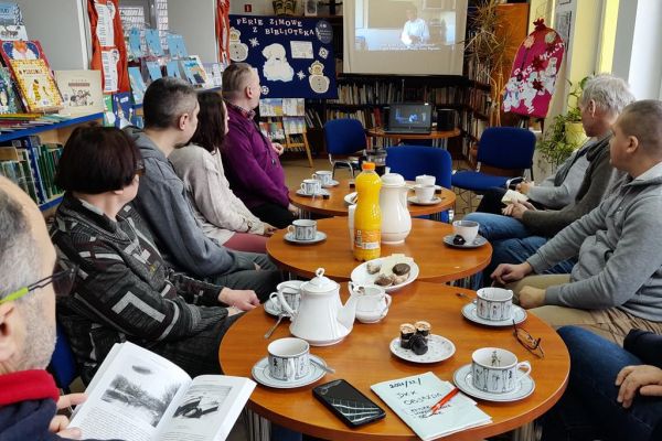 Mieszkańcy siedzą w bibliotece przy stolikach i wysłuchują Pani prowadzącej.