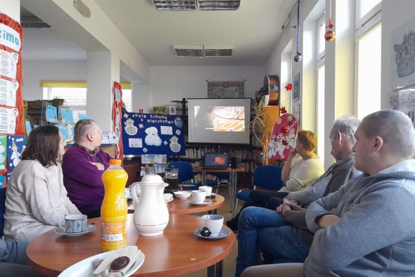 Mieszkańcy siedzą w bibliotece przy stolikach i wysłuchują Pani prowadzącej.