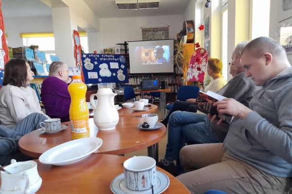 Mieszkańcy siedzą w bibliotece przy stolikach i wysłuchują Pani prowadzącej.