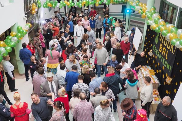 Uczestnicy balu bawiący się na holu domu udekorowanym złotymi, zielonymi i perłowymi balonami.