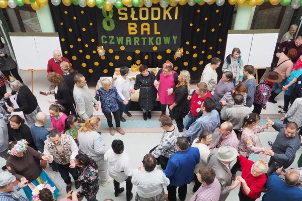 Uczestnicy balu bawiący się na holu domu udekorowanym złotymi, zielonymi i perłowymi balonami, na tle napisu na czarnej kotarze w złote kropki.