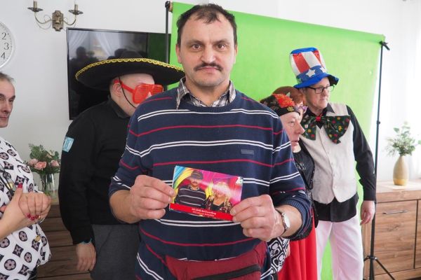 Uczestnik balu pokazujący zdjęcie z fotobudki a za nim troje uczestników w przebraniu pozuje do zdjęcia w fotobudce.
