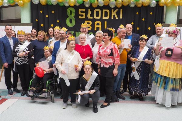 Nagrodzeni uczestnicy balu pozują do zdjęcia w szarfach i koronach, trzymając dyplomy, a z nimi stoją niektórzy Dyrektorowie domów na tle napisu na kotarze.