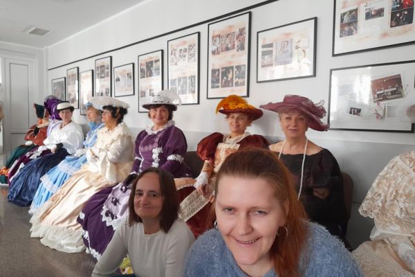 Dwie mieszkanki kucające przed rzędem siedzących Paniach w sukniach z XIX w.