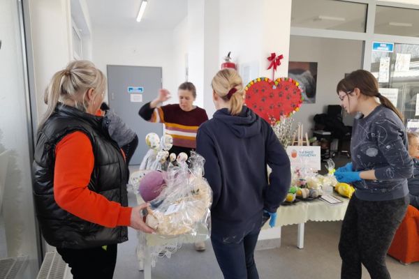 Pani z UTW Rędzikowo oferuje prace na kiermaszu wyłożone na stole w schronisku, a odwiedzający schronisko (tu trzy osoby) oglądają je.