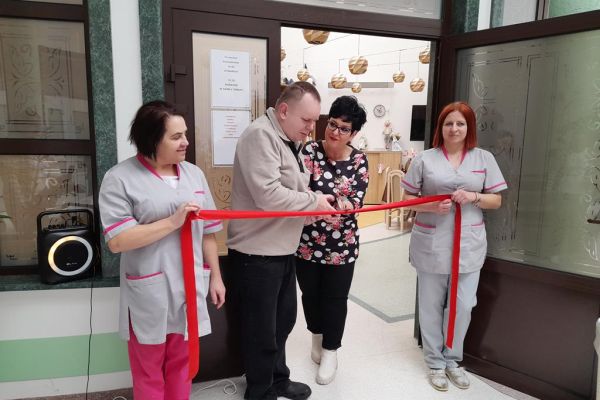 Mieszkaniec wraz z Panią Dyrektor przecina wstęgę na wejściu do nowej terapii trzymaną przez dwie Panie.