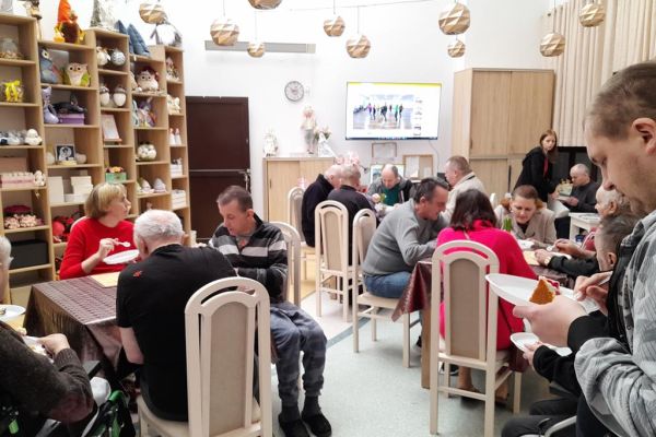 Mieszkańcy w nowej sali terapi degustują tort siedząc przy stolikach.