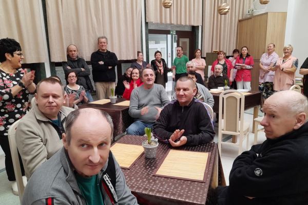 Mieszkańcy siedzą przy stolikach w nowej sali terapii i śpiewają karaoke wraz z pracownikami.