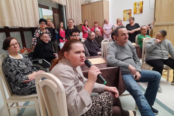 Mieszkańcy siedzą przy stolikach w nowej sali terapii i śpiewają karaoke wraz z pracownikami.