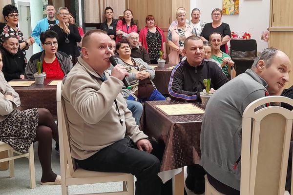 Mieszkańcy siedzą przy stolikach w nowej sali terapii i śpiewają karaoke wraz z pracownikami.