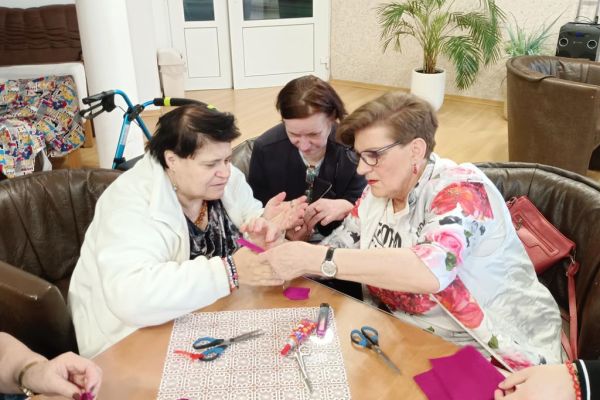 Mieszkańcy wraz z uczestnikami UTW z Redzikowa wykonują ozdoby siedząc przy stolikach w kawiarence na terenie DPS  w Lubuczewie.