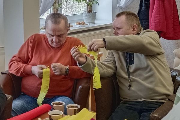 Mieszkańcy wraz z uczestnikami UTW z Redzikowa wykonują ozdoby siedząc przy stolikach w kawiarence na terenie DPS  w Lubuczewie.