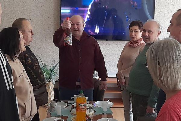 Kilku mieszkańców wraz z solenizantem świętują jego urodziny i imieniny przy stolikach w kawiarence i przy słodkościach i napojach oraz torcie.