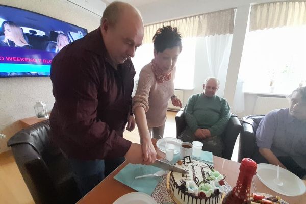 Kilku mieszkańców wraz z solenizantem świętują jego urodziny i imieniny przy stolikach w kawiarence i przy słodkościach i napojach oraz torcie.