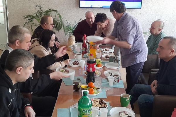 Kilku mieszkańców wraz z solenizantem świętują jego urodziny i imieniny przy stolikach w kawiarence i przy słodkościach i napojach oraz torcie.