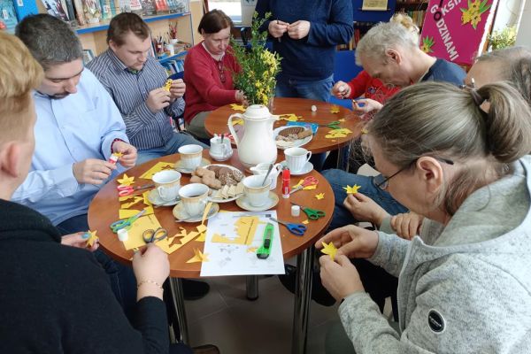 Mieszkańcy w bibliotece przy stolikach wycinają żółte żonkile z papieru..