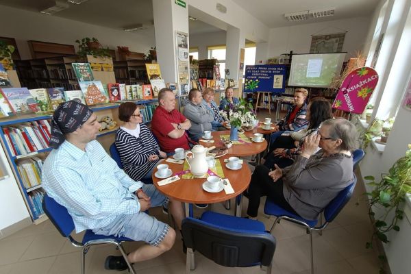 Mieszkańcy siedzą przy stolikach w bibliotece i uczestniczą w spotkaniu DKK.