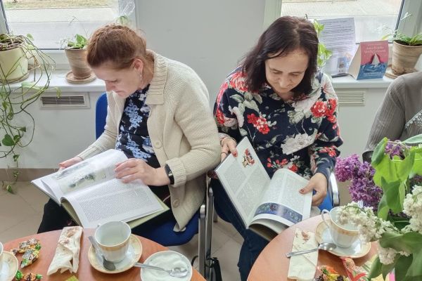 Mieszkańcy siedzą przy stolikach w bibliotece i uczestniczą w spotkaniu DKK oraz czytają książki..