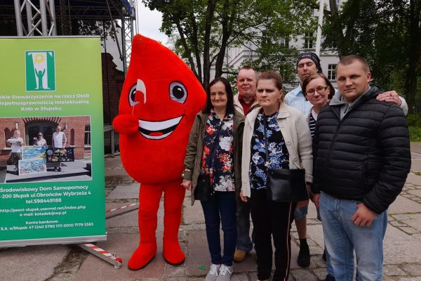 Mieszkańcy w parku w Słupsku podczas Dnia Godności Osób z Niepełnosprawnością Intelektualną stoją w gupowym zdjęciu z maskotką Kropli krwi.
