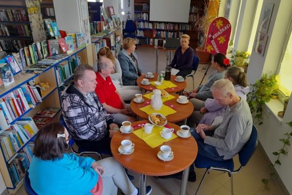 Mieszkańcy siedzą przy stolikach z poczęstunkiem w bibliotece na prelekcji.