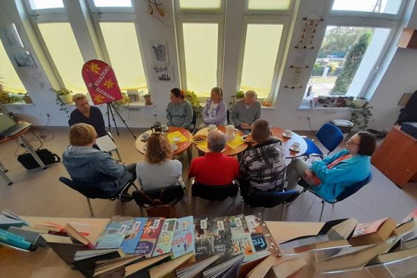 Mieszkańcy siedzą przy stolikach z poczęstunkiem w bibliotece na prelekcji.