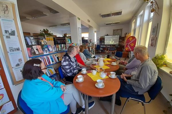 Mieszkańcy siedzą przy stolikach z poczęstunkiem w bibliotece na prelekcji.