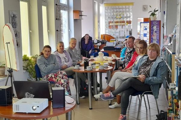 Mieszkańcy siedzą przy stolikach z poczęstunkiem w bibliotece na prelekcji.