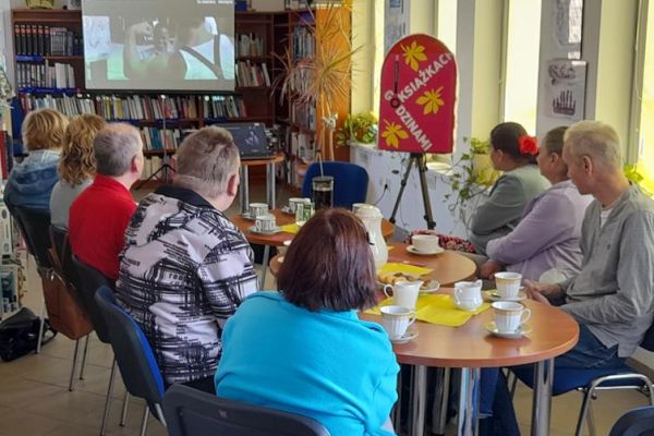 Mieszkańcy siedzą przy stolikach z poczęstunkiem w bibliotece na prelekcji.