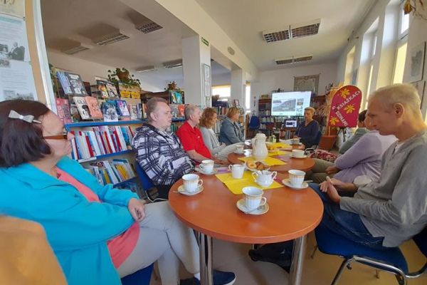 Mieszkańcy siedzą przy stolikach z poczęstunkiem w bibliotece na prelekcji.
