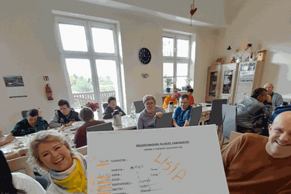 Mieszkańcy wraz z innymi uczestnikami projektu przy stolikach z poczęstunkiem., a na pierwszym planie lizta z podziałem na role w sztyce teatralnej.
