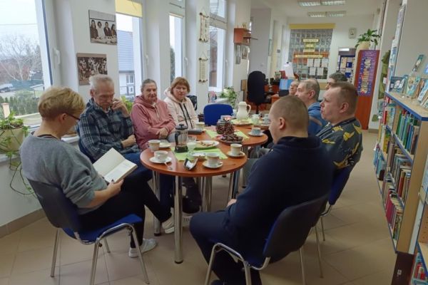 Mieszkańcy słuchaja opowiadania siedząc przy stolikach w bibliotece.