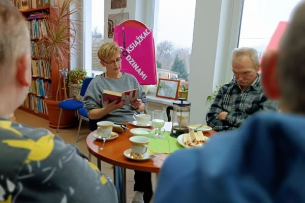 Mieszkańcy słuchaja opowiadania siedząc przy stolikach w bibliotece.