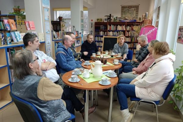 Mieszkańcy słuchaja opowiadania siedząc przy stolikach w bibliotece.