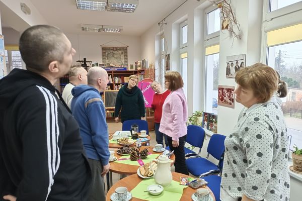 Mieszkańcy stoją przy stolikach w bibliotece i wykonują ćwiczenia rozluźniające.